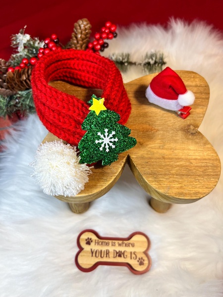 A close-up, high-quality product shot of a red knitted dog scarf featuring a sparkly green Christmas tree applique, displayed on a wooden stand with a wooden bone sign.