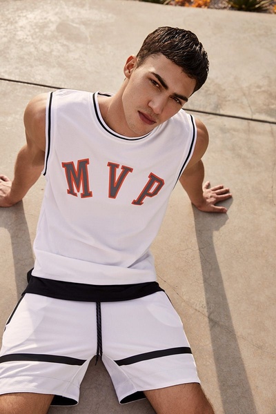 A high-quality lifestyle photograph of a male model wearing a branded athletic tank top and shorts, shot from a high angle in a sunlit outdoor setting.