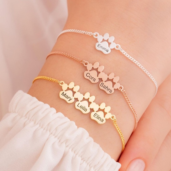A close-up, warm-toned image showing three variations of a delicate name-engraved paw bracelet being worn on a wrist.