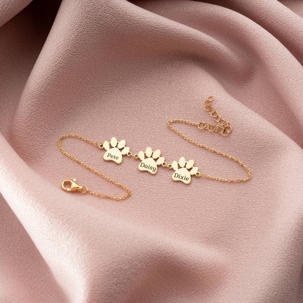 A high-quality studio shot of a personalized gold paw-print bracelet resting on soft, flowing pink fabric to evoke an emotional connection.