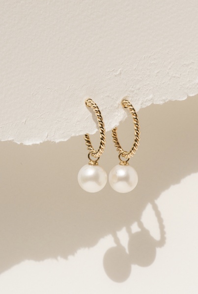 A close-up, soft-focus shot of gold twisted-hoop earrings with pearl drops hanging from a textured paper edge.