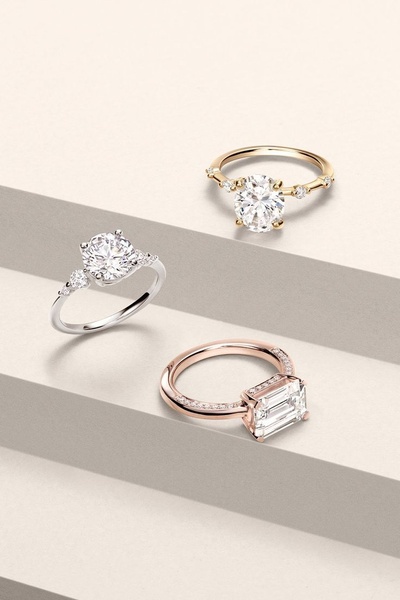 A high-end studio display featuring three engagement rings in silver, gold, and rose gold finishes on a stepped neutral platform.