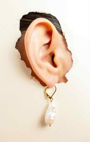 A close-up photograph of a pearl earring worn on an ear, emerging from a tear in a textured, light-colored background.