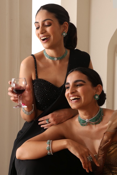 Two models showcasing emerald necklace and jewelry sets in a sophisticated social setting with dark aesthetic branding.