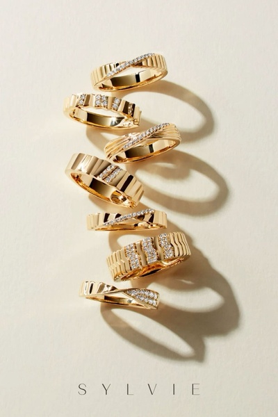 A vertical composition of multiple gold rings with diamond accents arranged on a soft, neutral, textured background.