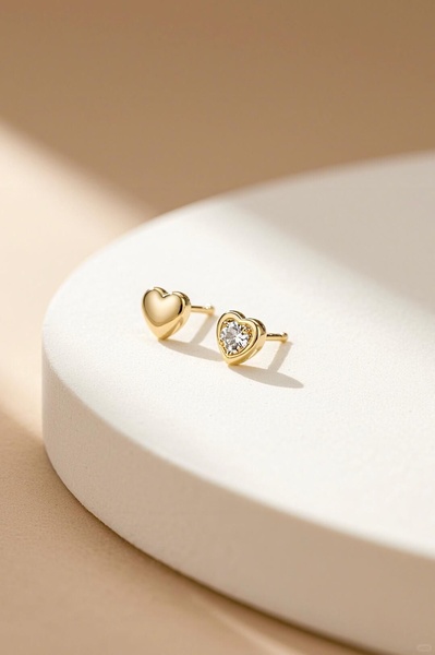 A refined, high-end product hero shot featuring gold heart-shaped earrings displayed on a clean, light-colored pedestal with soft directional lighting.