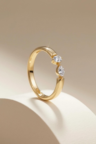A close-up studio shot of a delicate gold ring featuring two clear stones on a neutral, curved pedestal.