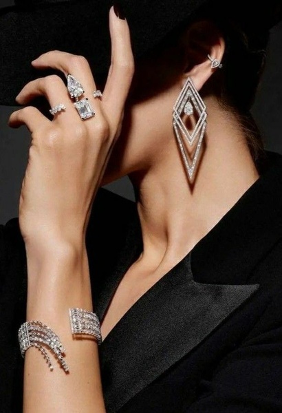 A close-up, high-fashion shot featuring a model wearing diamond earrings, bracelets, and rings against a dark, minimalist background.