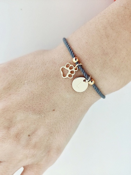 A close-up photograph of a minimalist gray cord bracelet featuring a gold paw print charm and an initial disc, displayed on a wrist.