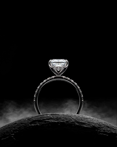A high-contrast, moody close-up of a diamond engagement ring perched on a dark, textured stone base, surrounded by atmospheric light.