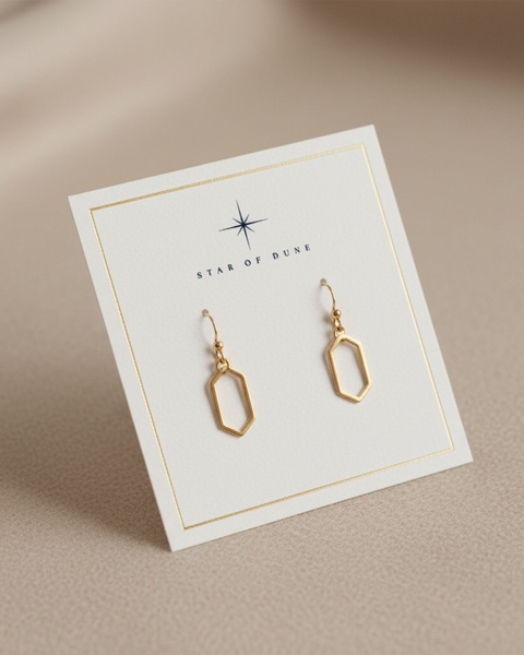 A close-up studio shot of a pair of gold drop earrings displayed on a branded white card against a soft, neutral beige backdrop.