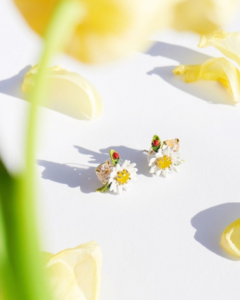 A close-up, high-key photograph of two daisy-themed earrings placed on a white surface, surrounded by yellow tulip petals.