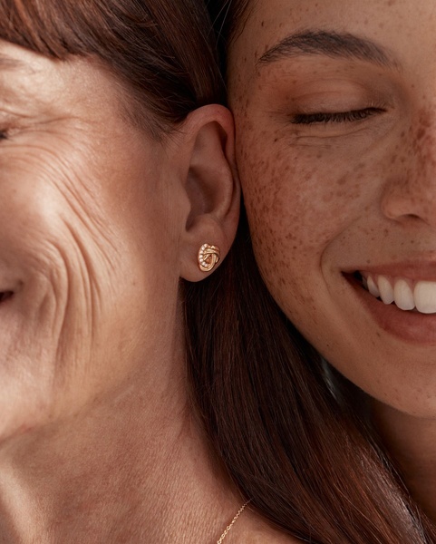 A close-up photograph featuring two women in a warm, intimate embrace highlighting a gold knot-style earring.
