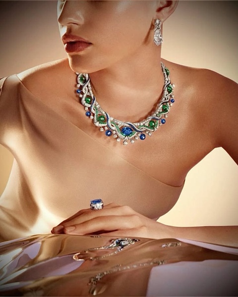 A close-up portrait of a model wearing an intricate blue, green, and silver necklace with matching jewelry, set against a warm, minimalist studio background.