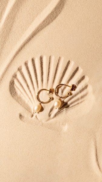 A high-contrast overhead shot of gold hoop earrings with pearls resting on a natural sand seashell imprint.