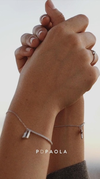 A close-up, high-contrast shot of two hands holding each other, highlighting matching delicate chain bracelets with charm accents.