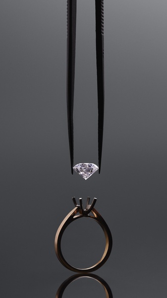 A high-end, studio-lit creative showing a diamond being placed into a ring setting using precision tweezers against a clean grey backdrop.
