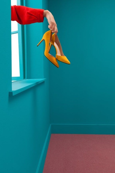 A striking visual display of a pair of yellow high-heels being held by a hand through a window against a bold teal background.