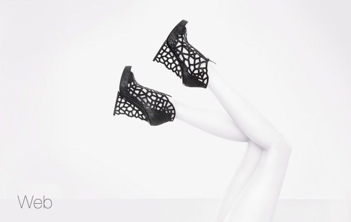 A high-fashion footwear advertisement featuring a pair of black geometric wedges held up by white mannequin-style legs against a stark white background.