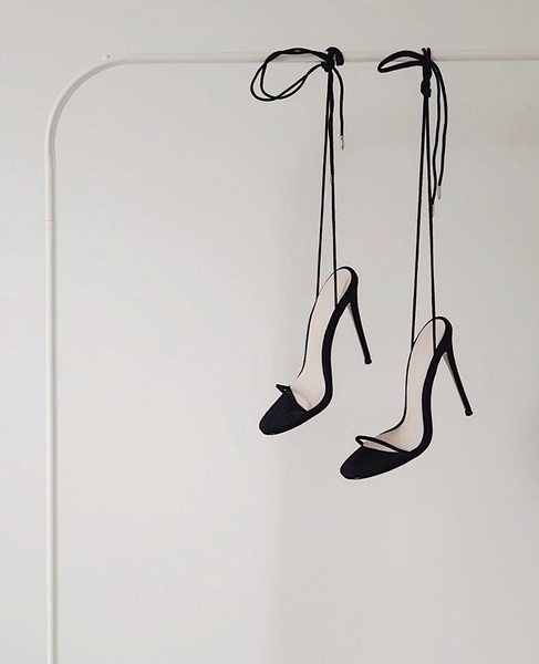A high-end, clean studio shot of black strapped high heels hanging from a minimalist white rack against a plain backdrop.