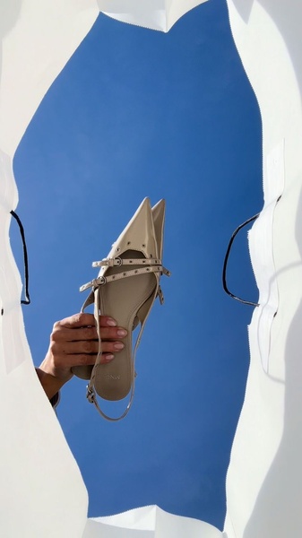 A high-angle, minimalist photograph featuring a pair of neutral-toned pointed toe flats held against a bright blue sky.