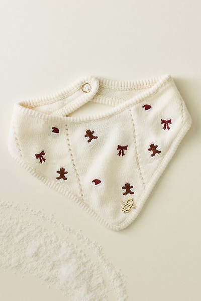 A close-up studio shot of a cream-colored knit baby bib featuring embroidered gingerbread men, ribbons, and Santa hats on a neutral background.