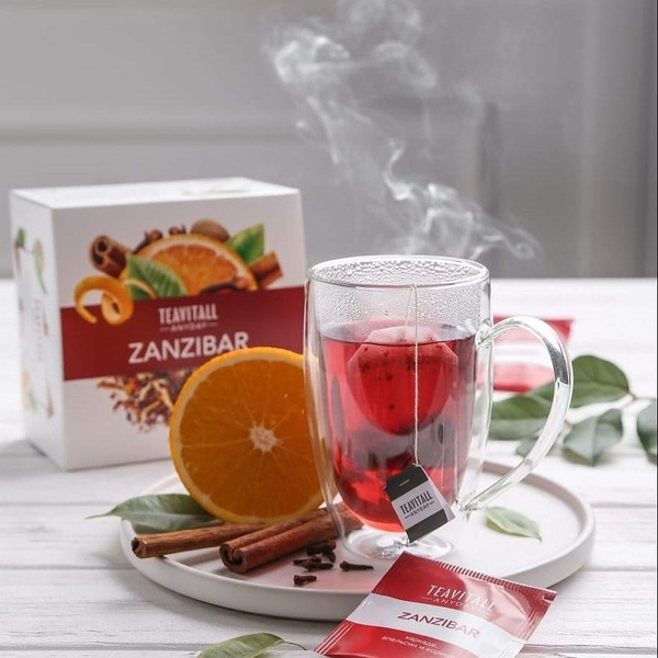A close-up, high-definition photograph of a steaming cup of fruit tea served on a white tray, surrounded by oranges, cinnamon sticks, and cloves.