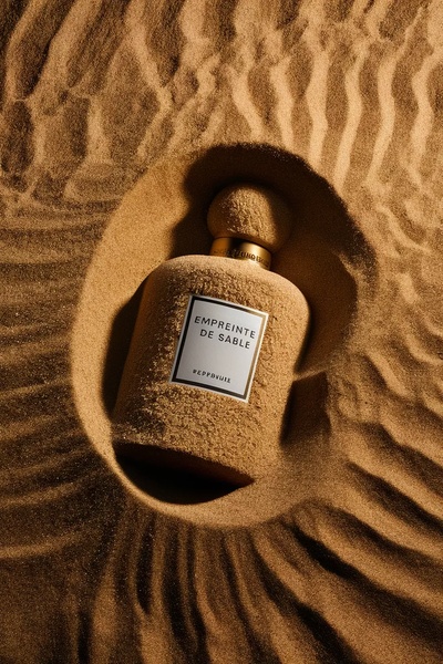 A high-end studio shot of a perfume bottle partially buried in fine, textured desert sand, highlighting a luxury minimalist aesthetic.