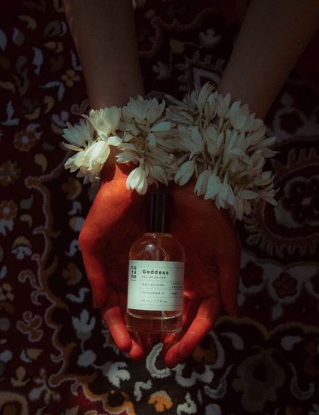 A close-up aesthetic shot of hands holding a clear perfume bottle adorned with white flowers, set against a dark patterned rug.