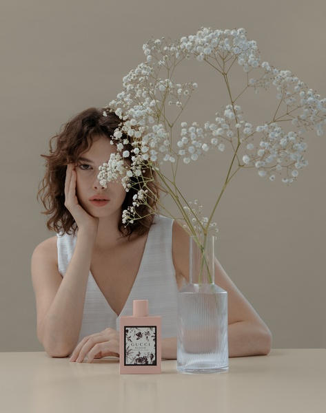 A refined product shot featuring a perfume bottle placed on a table next to a vase of baby's breath, with a model's face partially obscured by the flowers to create a sense of mystery and elegance.