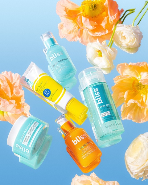 A flat-lay inspired arrangement of various skincare bottles and jars suspended against a vibrant blue sky with scattered floral accents.