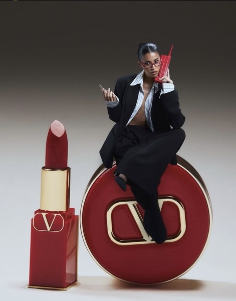 A high-fashion, studio-shot editorial creative featuring a lipstick positioned next to a giant, stylized version of the product casing and a model on a phone.