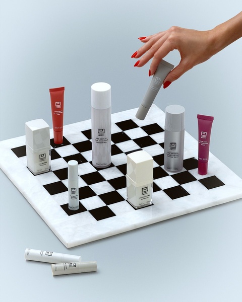 A high-end skincare collection is neatly organized on a marble chessboard, with a hand selecting a product tube, embodying a luxury aesthetic.