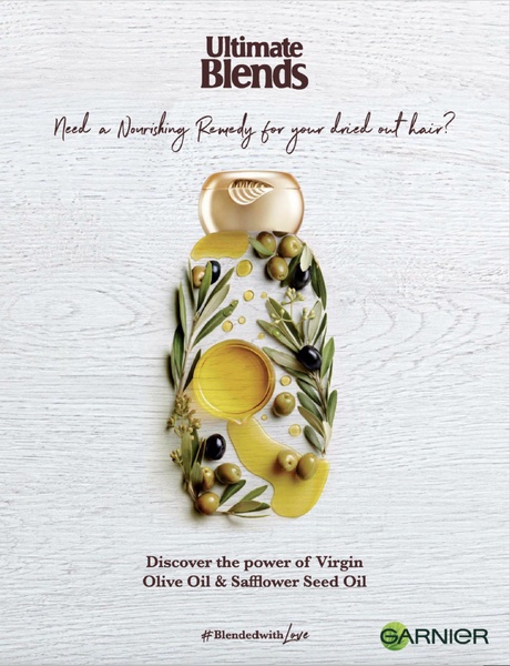 This ad creative uses a clever layout where olive oil, herbs, and leaves are arranged to form the silhouette of a shampoo bottle against a rustic, white-washed wooden background.