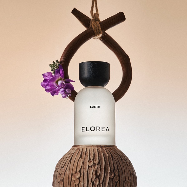 A minimalist, high-end studio shot of a glass perfume bottle suspended within a wooden ring, decorated with a small cluster of purple flowers.