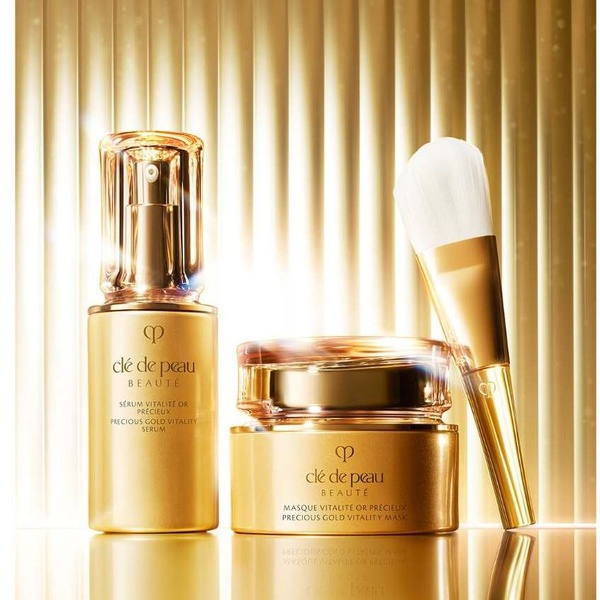 A high-end, studio-lit photograph showcasing a gold serum bottle, a matching cream jar, and an applicator brush against a ridged metallic background.