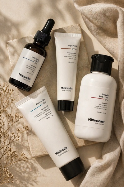A high-quality, top-down flat lay composition featuring four essential skincare bottles on a textured stone platform surrounded by neutral linens.