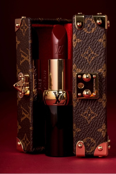 A close-up studio photograph of a luxury lipstick presented inside a miniature trunk, emphasizing premium craftsmanship and rich, deep color tones.