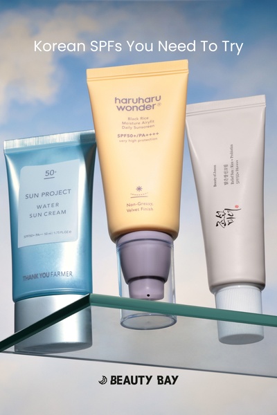 A flat lay studio shot showcasing three different Korean sunscreens arranged on a glass shelf against a soft, blue sky background.