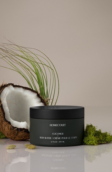 A high-end, clean product shot featuring body butter centered against a neutral background with organic coconut and moss accents.