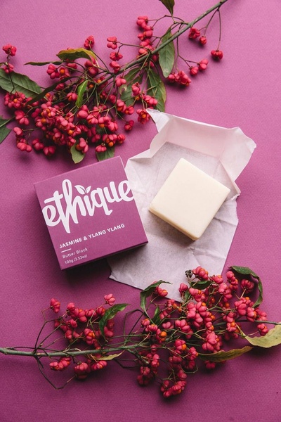 A top-down, minimalist product shot featuring a solid butter block centered on a vibrant purple background surrounded by floral botanical accents.