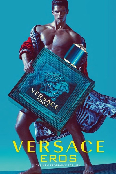 A high-fashion, high-contrast studio portrait featuring a male model showcasing a large fragrance bottle against a vibrant blue backdrop.