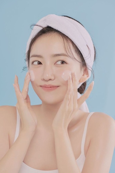 A clean, high-key studio shot of a model applying skin care product to her cheeks against a soft blue background.