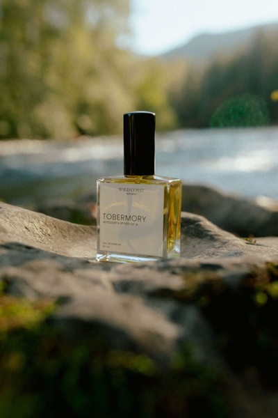A high-quality close-up shot of a Tobermory perfume bottle resting on a natural, mossy stone in front of a blurred river scene.