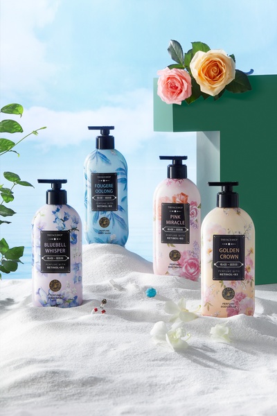 A high-end editorial display of four perfume body wash bottles nestled in white sand with fresh flowers and a soft sky-blue background.