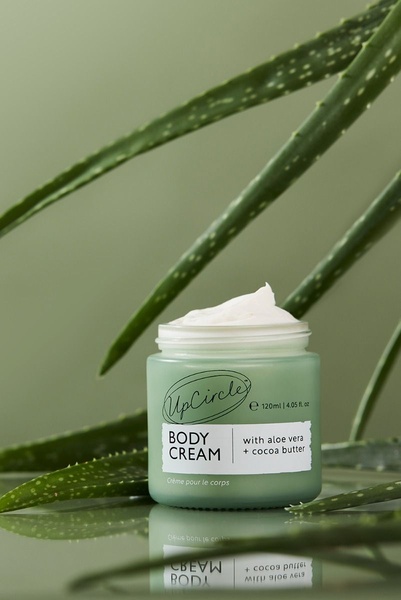 A clean, high-quality product shot featuring body cream inside a glass jar surrounded by fresh aloe vera leaves on a reflective surface.