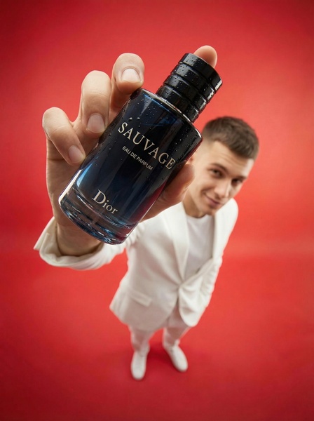 A close-up shot of a cologne bottle held by an individual in a white suit, using an exaggerated wide-angle perspective against a solid red background.