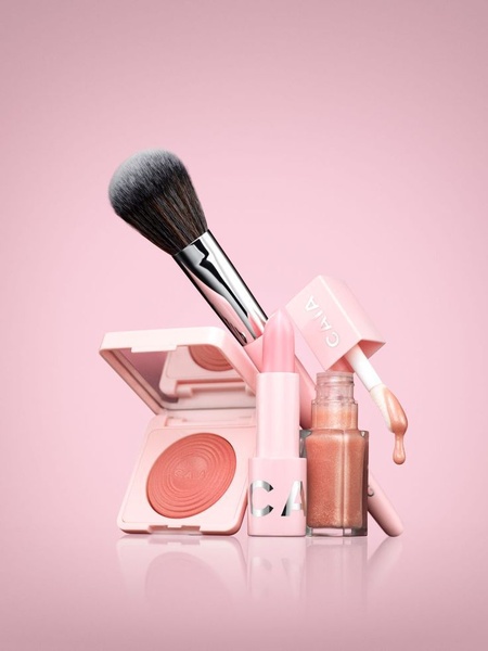 A clean, studio-lit arrangement of cosmetics products including a blush compact, lipstick, and liquid product against a solid pink backdrop.