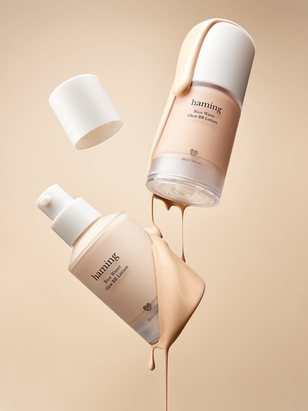 Two bottles of bb lotion are suspended in mid-air with creamy product pouring from the caps against a neutral, beige background.
