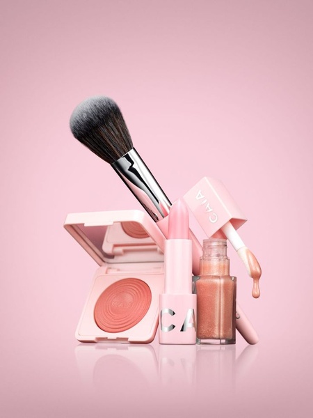 A clean, high-end studio photograph of a cosmetic set, including a brush, blush compact, lipstick, and lip gloss against a monochromatic pink background.
